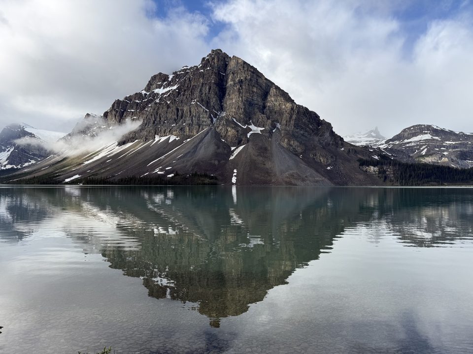 A rocky mountain reflected in a lake, with snow on its slopes and low cloud along the shoreline.